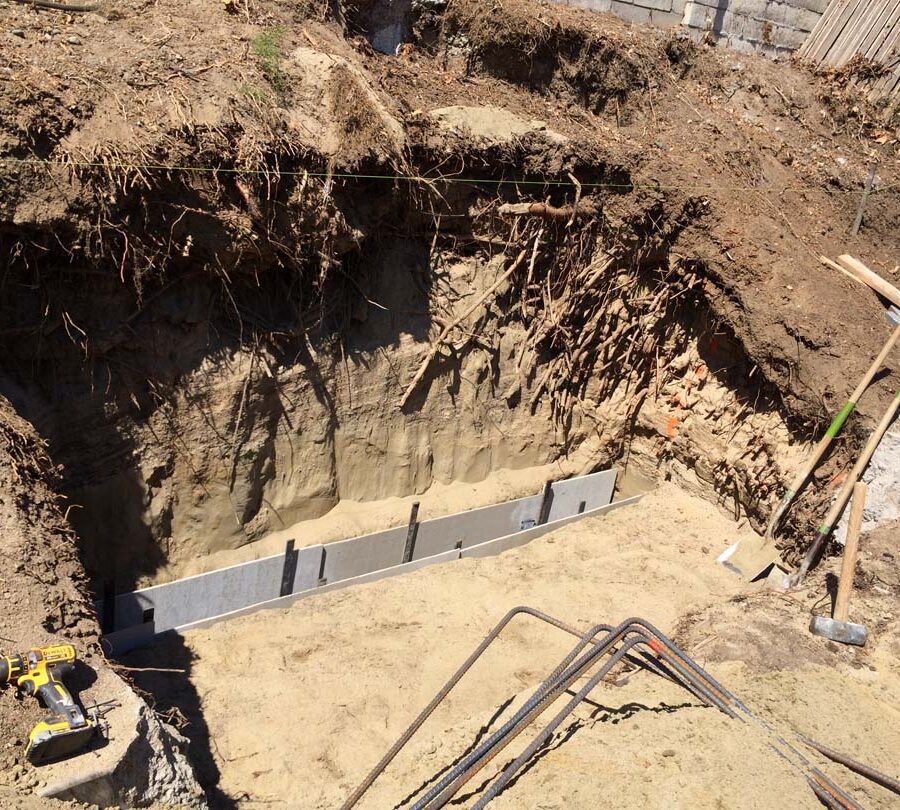 Excavating the strip footings for a new section of concrete retaining wall. This must be done in sections to avoid having the uneven soil collapse. Forms are temporarily placed to reinforce the existing sloped grade level. Pacificland Constructors